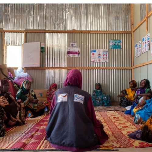 Eine Gruppe von Frauen in Somaliland nehmen an einer Gruppensitzung teil. Eine Gruppe von Frauen sitzen am Boden in einem Kreis. Im Vordergrund sieht man eine HI-Mitarbeiterin von hinten.